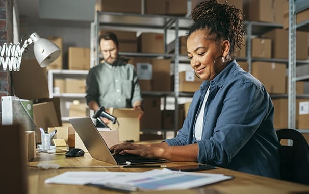 A woman is at a desk working at a laptop. A man is in the background, packing a shipping box.