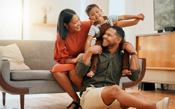 A man is seated on the floor in front of the sofa. His young child is seated on his shoulders, and his wife is seated on the sofa, helping to hold the child up. The family appears to be having fun and are happy.