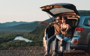A man and woman sit on the back of their car with the trunk open, staring at a scenic view
